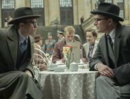 Zwei junge Männer mit Anzug und Krempenhut sitzen sich dicht nach vorne gebeugt an einem Café-Tisch gegenüber.